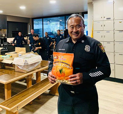 A police officer smiling while holding a bag of snacks in a kitchen.