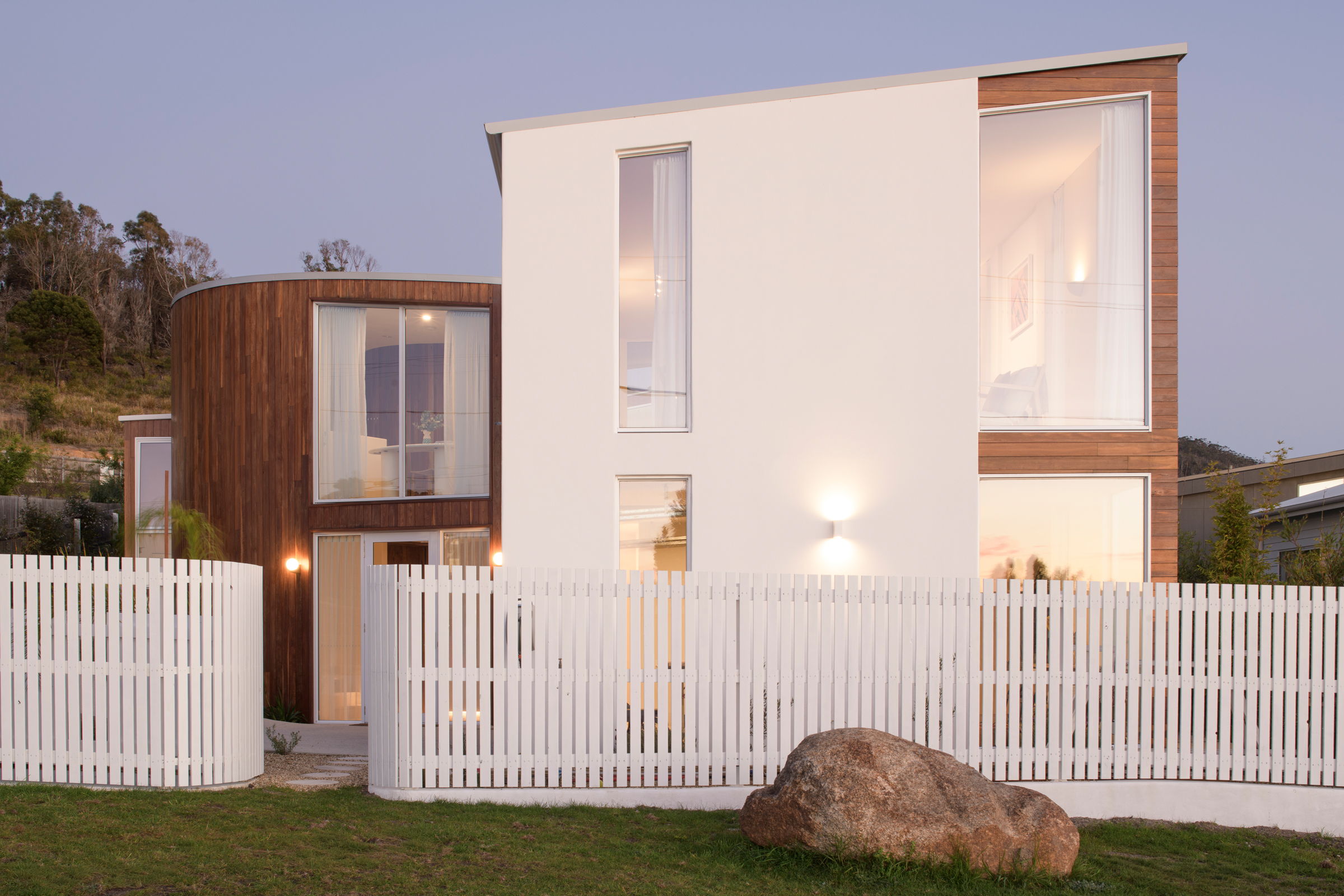 Modern house with wooden accents and white fence, surrounded by greenery.