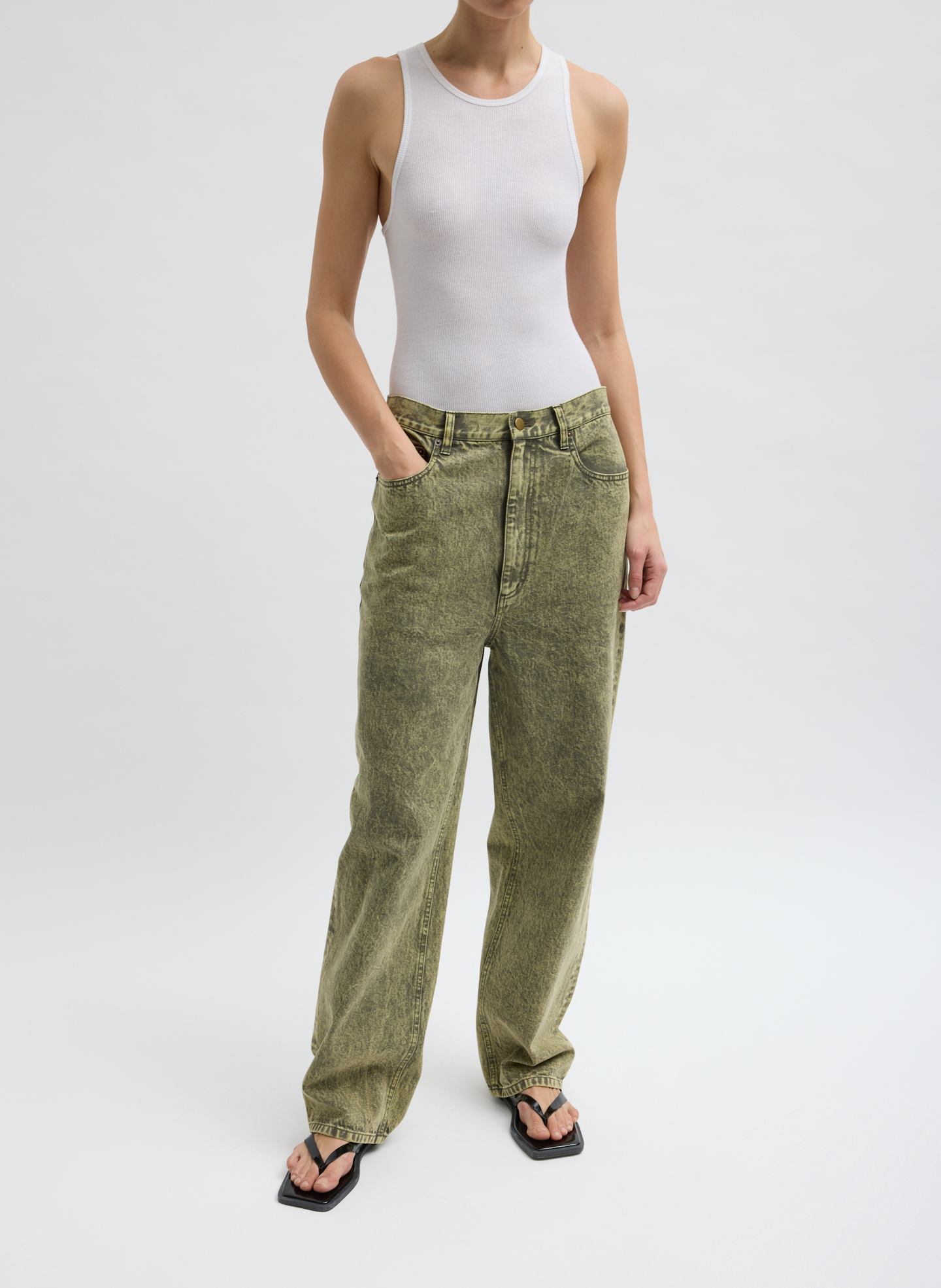 A person wearing a white tank top and green pants, standing with hands in pockets.