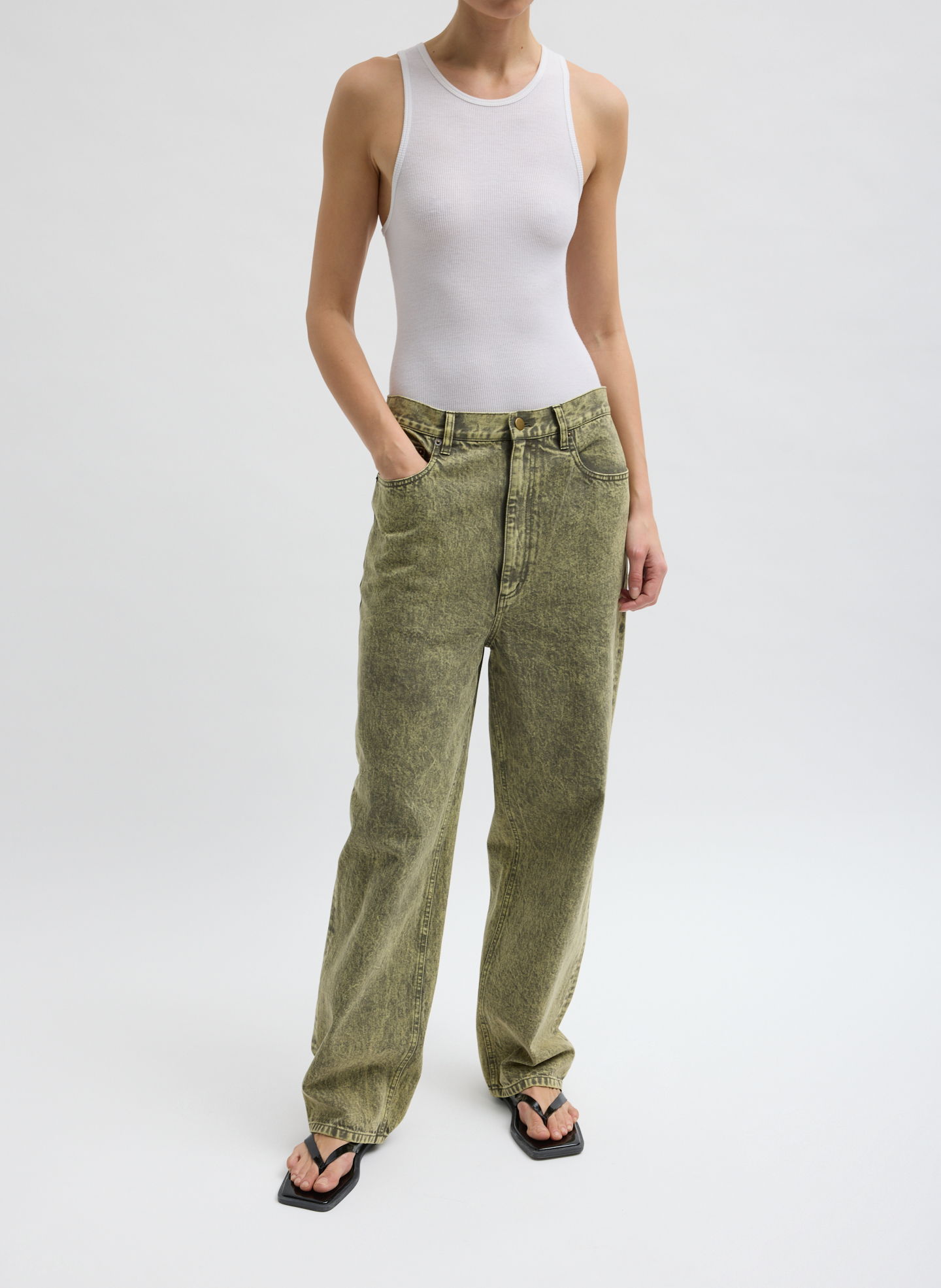 A person wearing a white tank top and green pants, standing with hands in pockets.