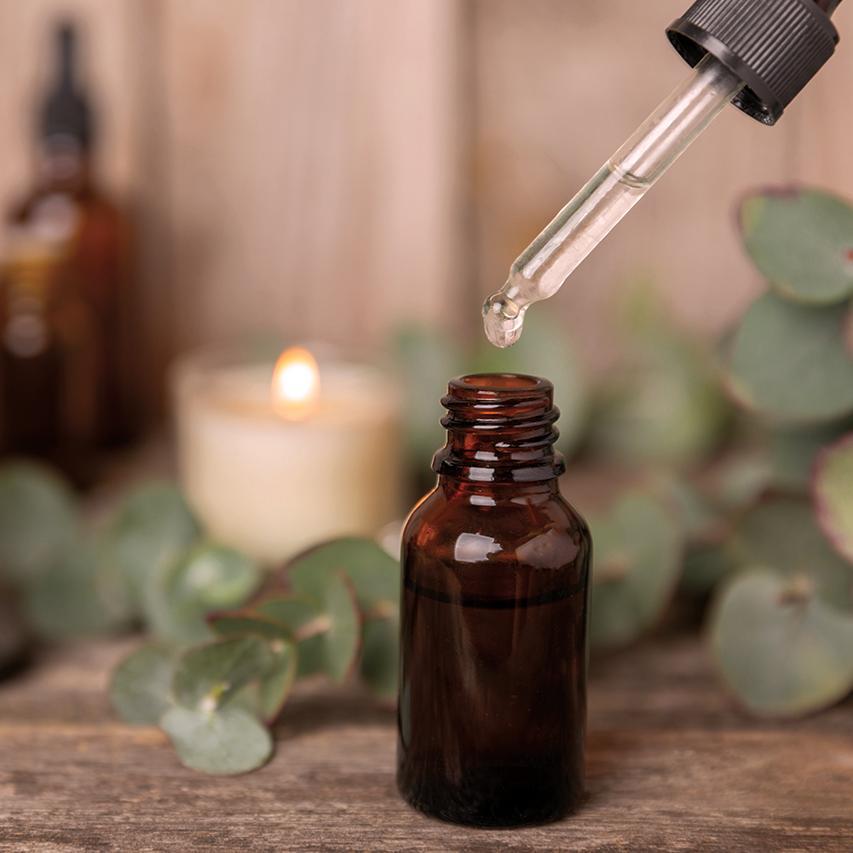 Dropper dispensing liquid into an amber bottle with candles and greenery.