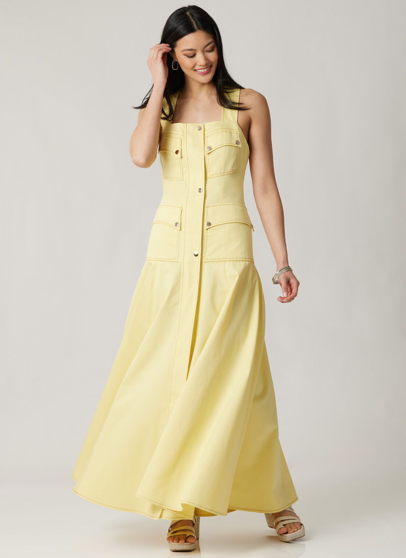 A smiling woman wearing a yellow maxi dress, standing and looking down.