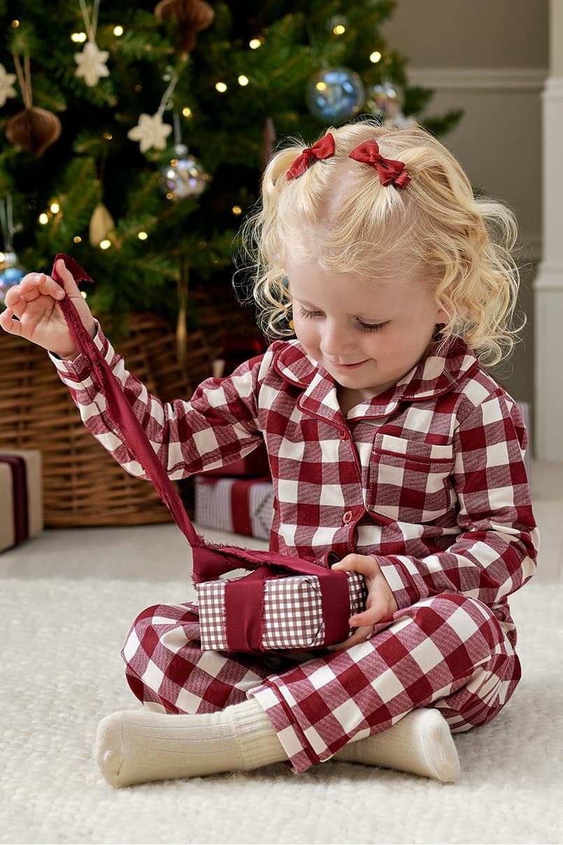 Child in pajamas opening a gift by a Christmas tree.