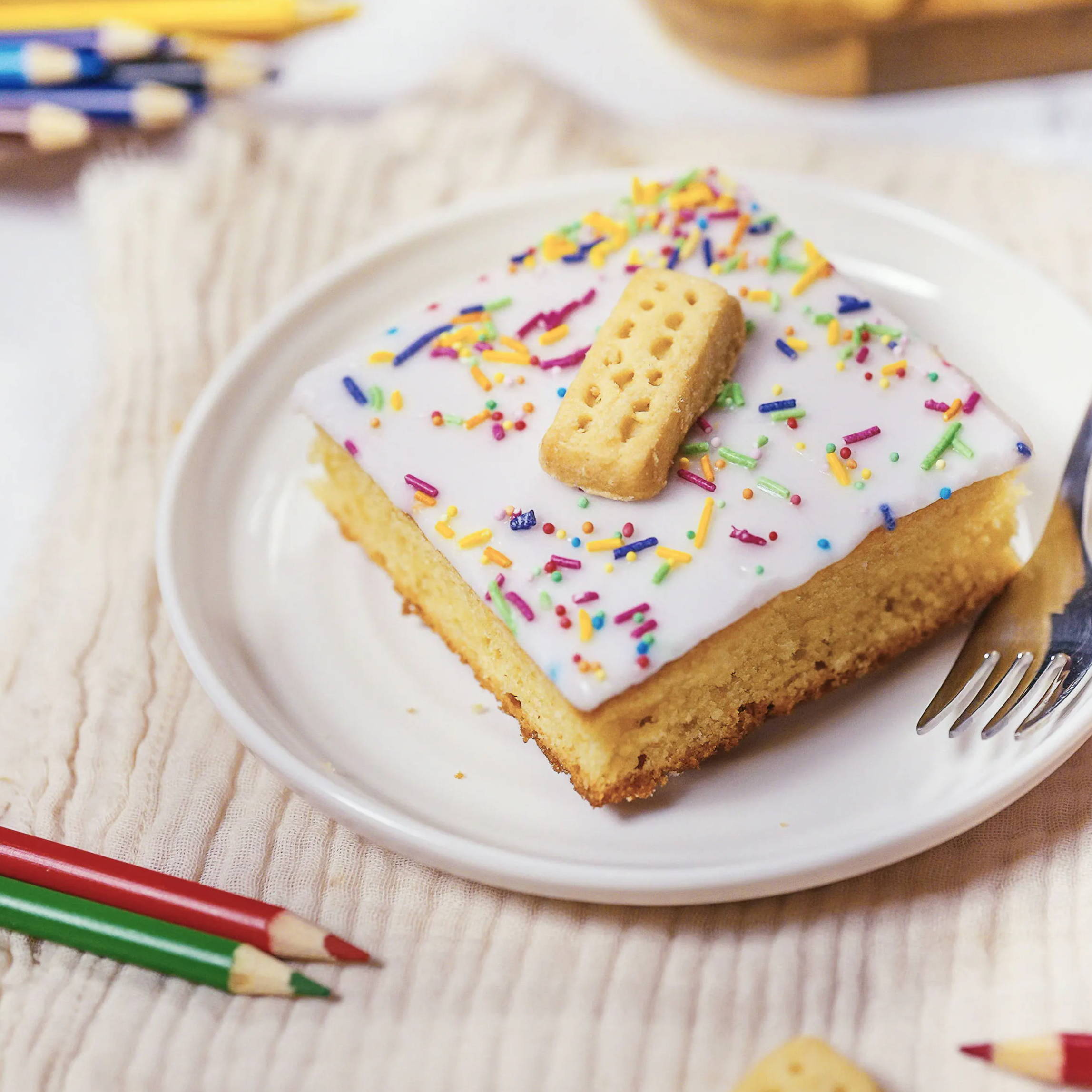 A slice of cake with frosting, sprinkles, and a biscuit on a plate.