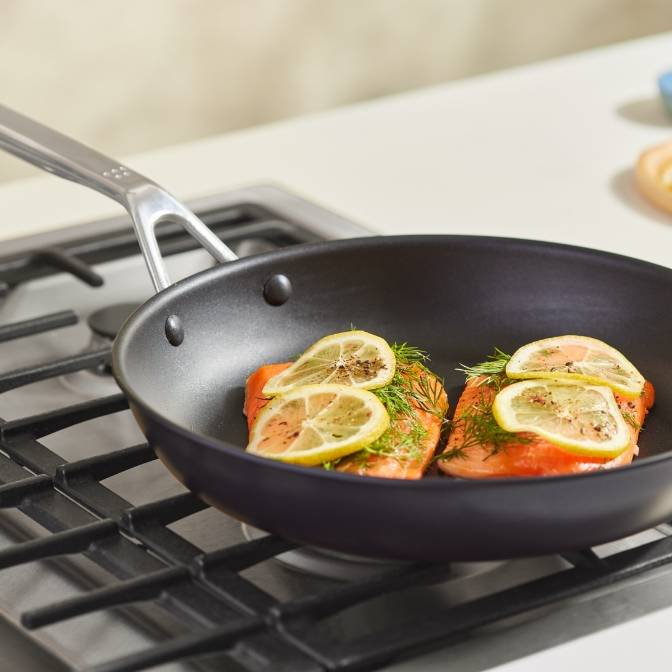 Two salmon filets topped with herbs and sliced lemon sit in a Misen Nonstick Pan on a stovetop.
