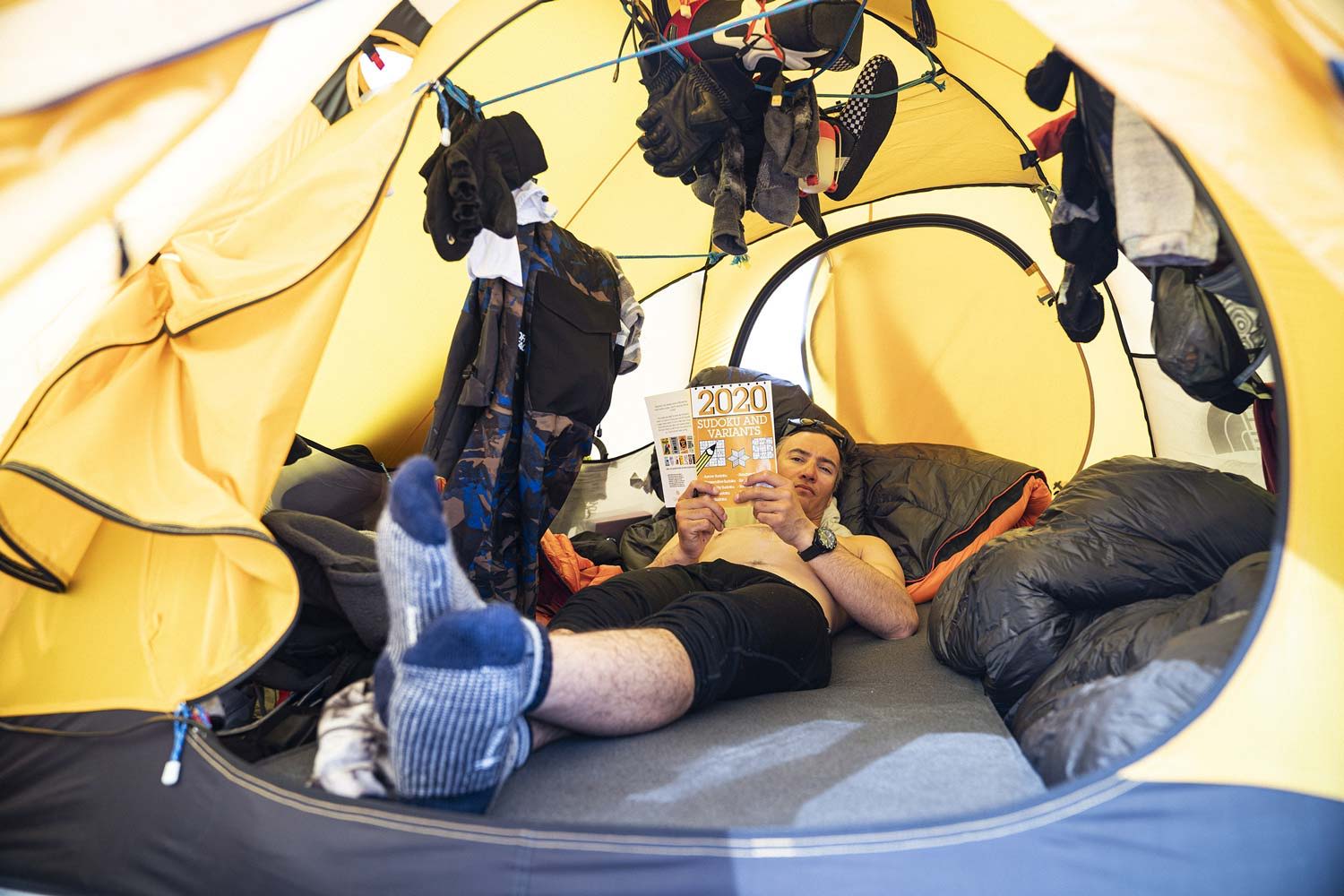 Person lounging inside a tent surrounded by gear and sleeping bags.