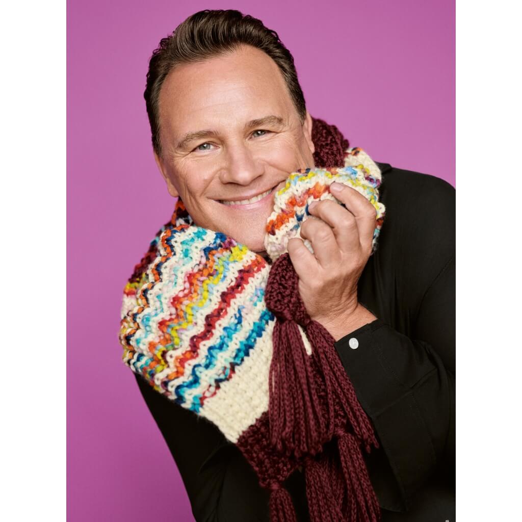 Eine lächelnde Person mit einem bunten Strickschal vor einem rosa Hintergrund.