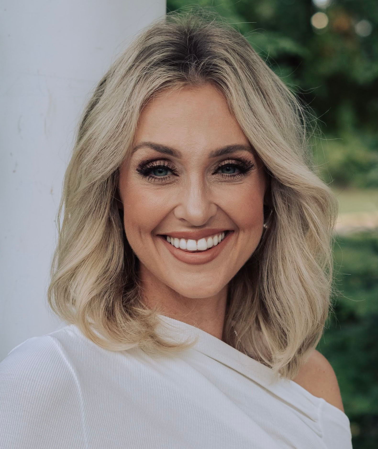 Head-and-shoulders portrait of a smiling woman with shoulder-length blonde hair wearing a white one-shoulder top. She is looking directly at the camera outdoors, with a softly blurred background of green trees and natural light.