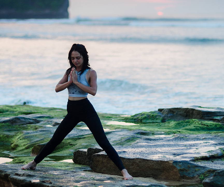 A woman in leggings and a sleeveless shirt standing on rocks at the beach in a meditative yoga pose.