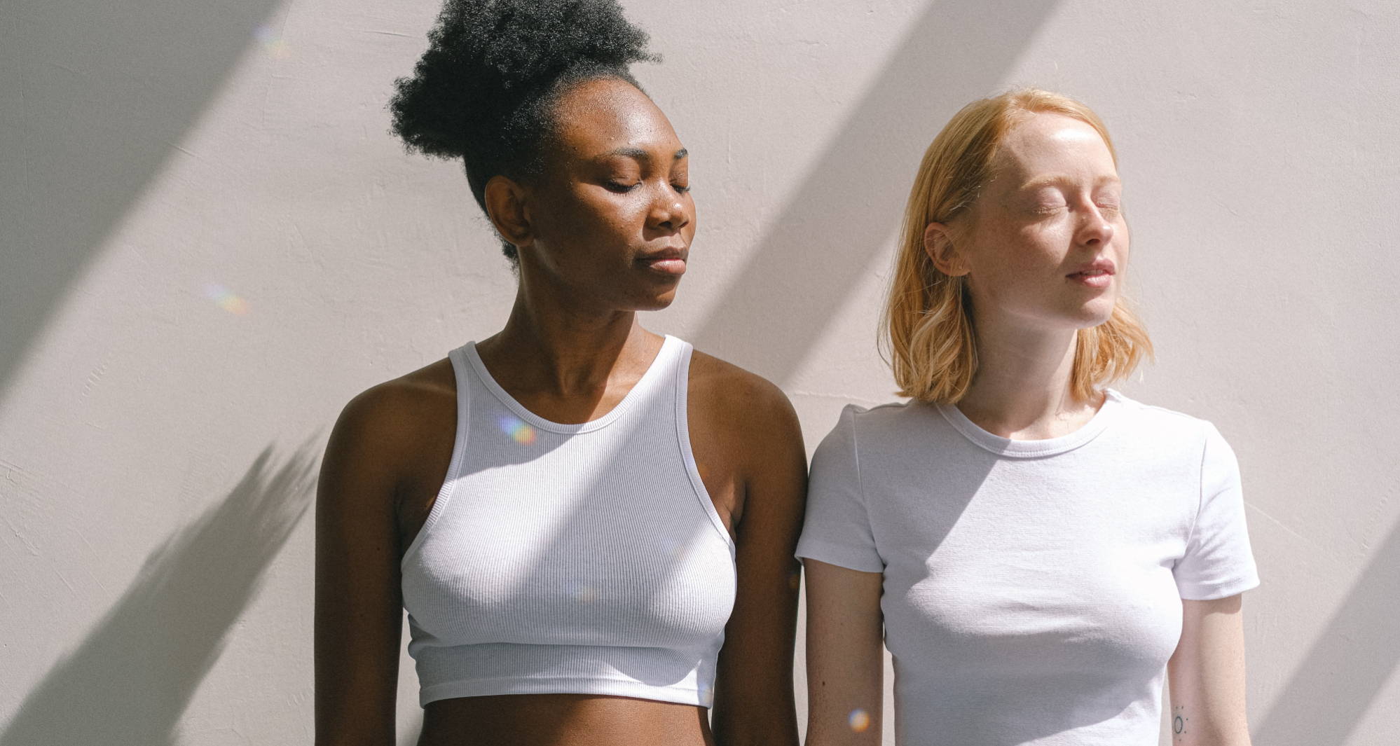  Two women standing in the sunshine with their eyes closed wear tight shirts without a visible bra.