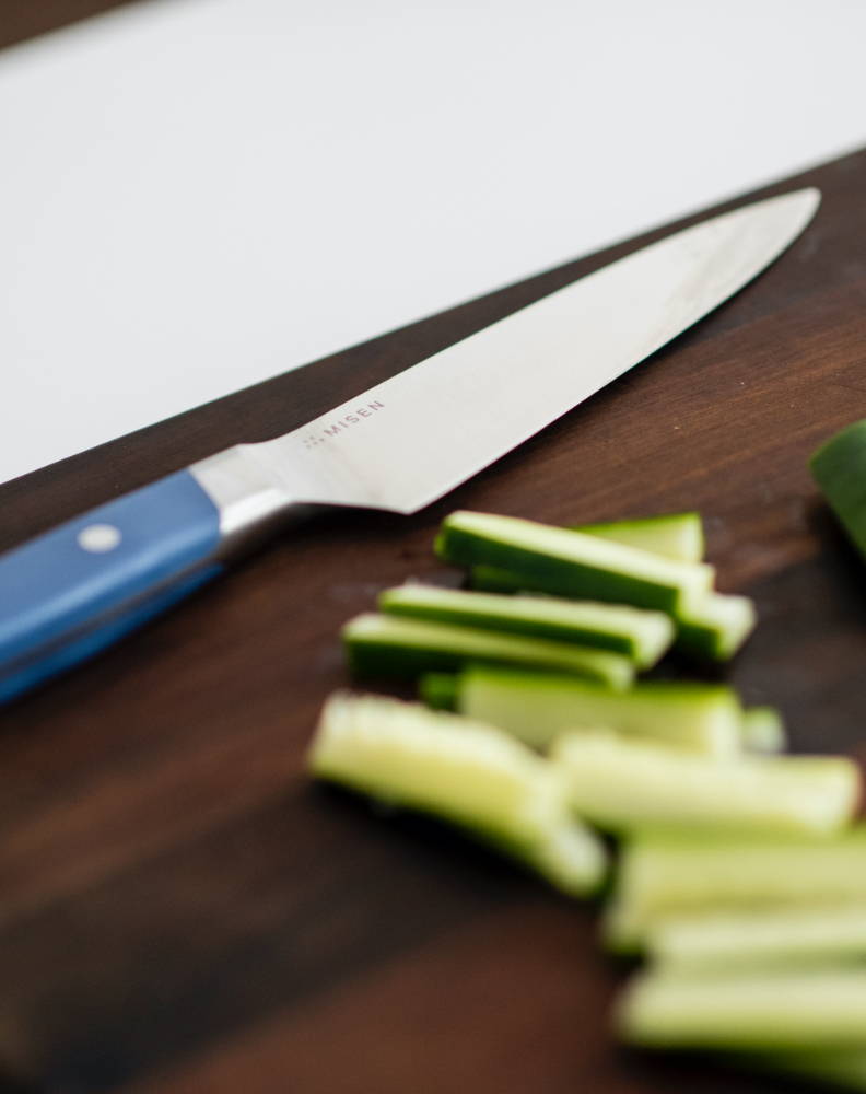 A Blue Misen Utility Knife resting on a dark wood cutting board, next to thinly-sliced cucumber sticks.