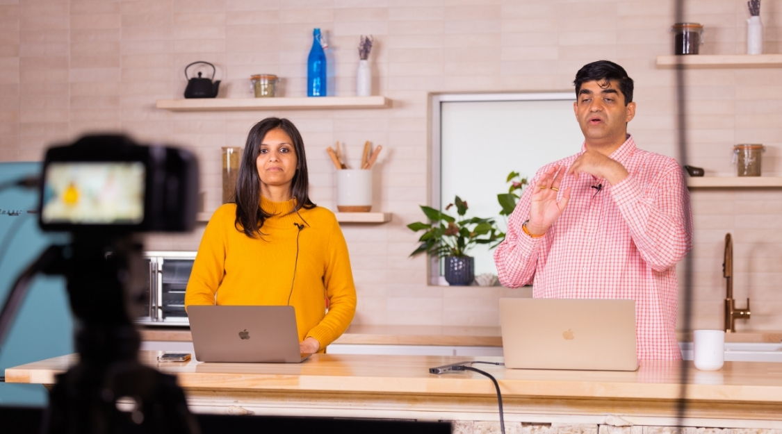 Two presenters in a kitchen setting, discussing with laptops and a camera.