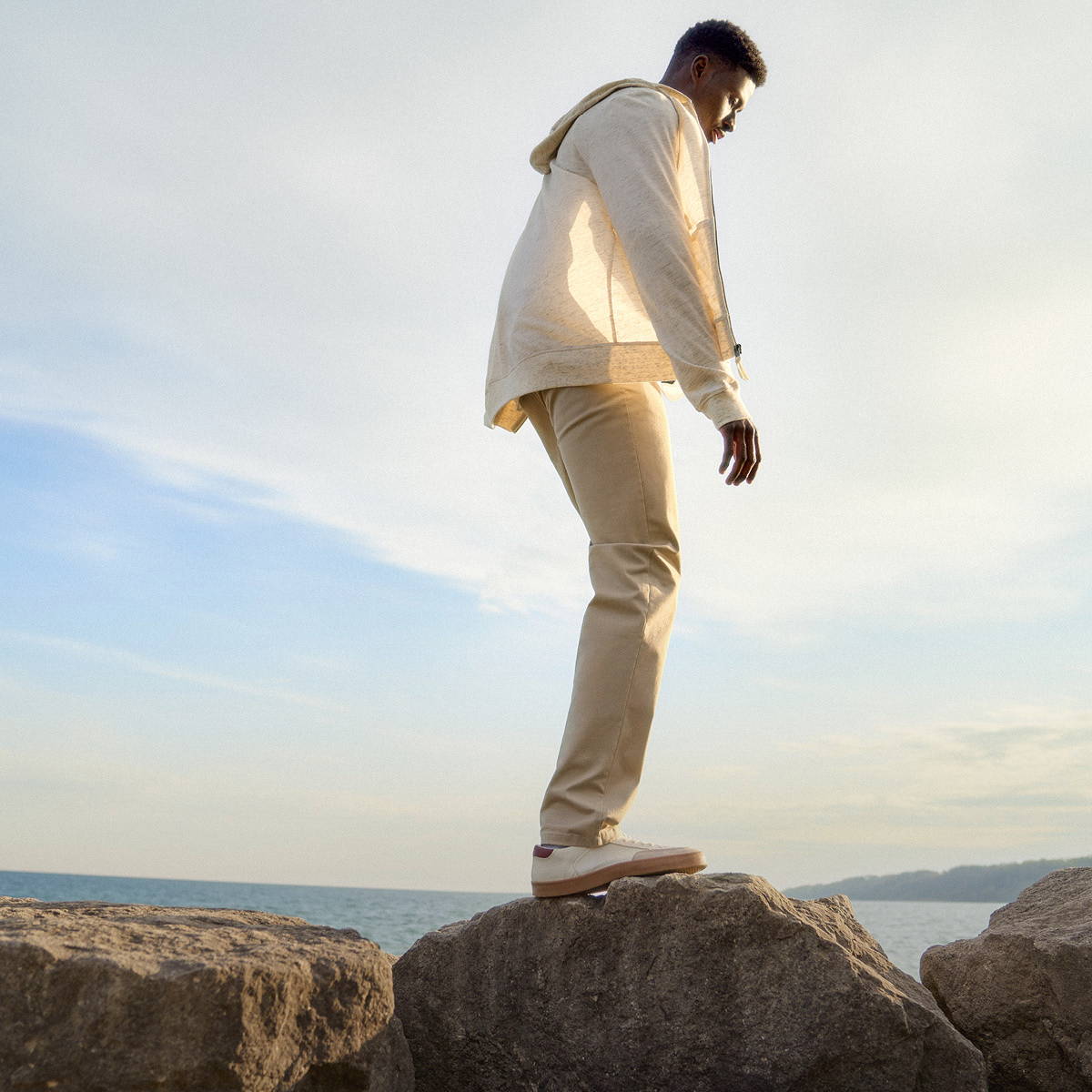 Tall man walking on rocks by the water wearing a beige sweater and khaki pants