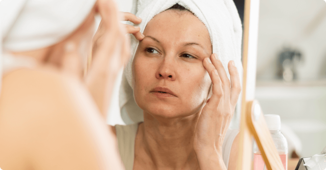 Woman with a towel on her head applying skincare in front of a mirror.