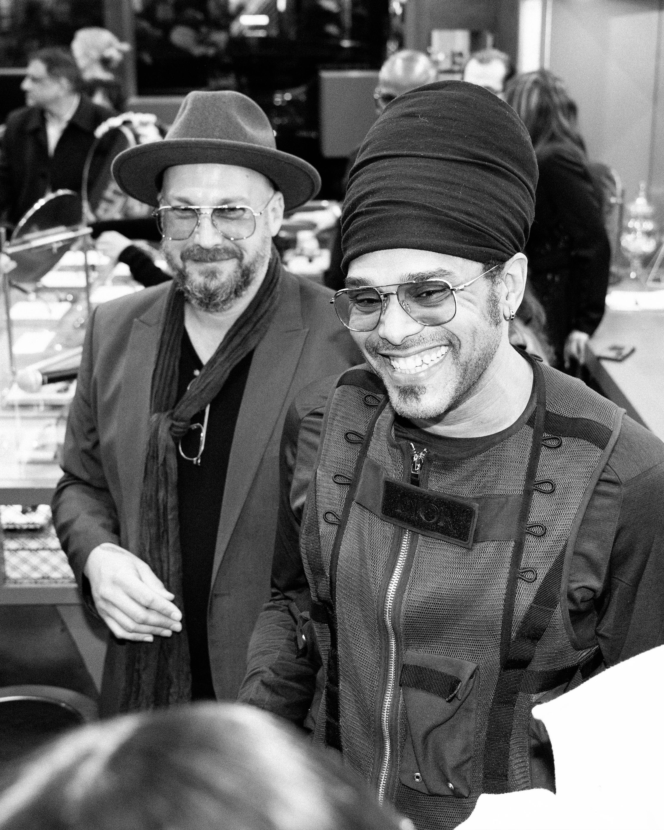Two men smiling, wearing stylish hats and glasses in a lively setting.