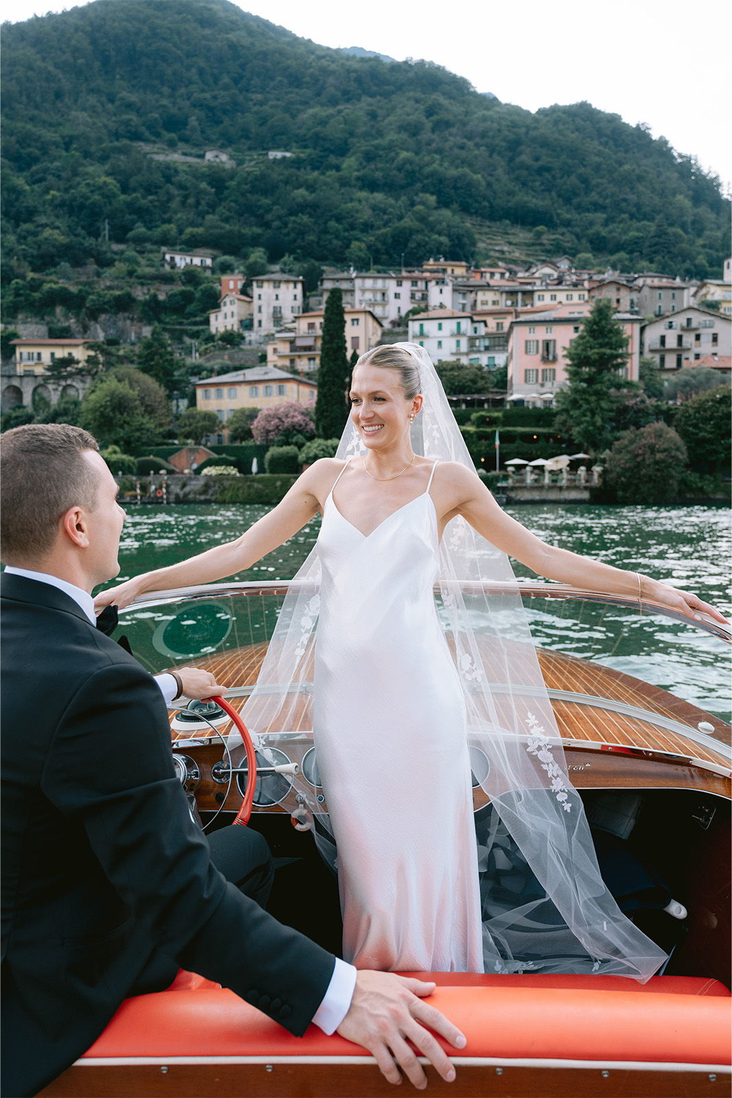 Bride in white dress on boat, smiling at groom with scenic backdrop.