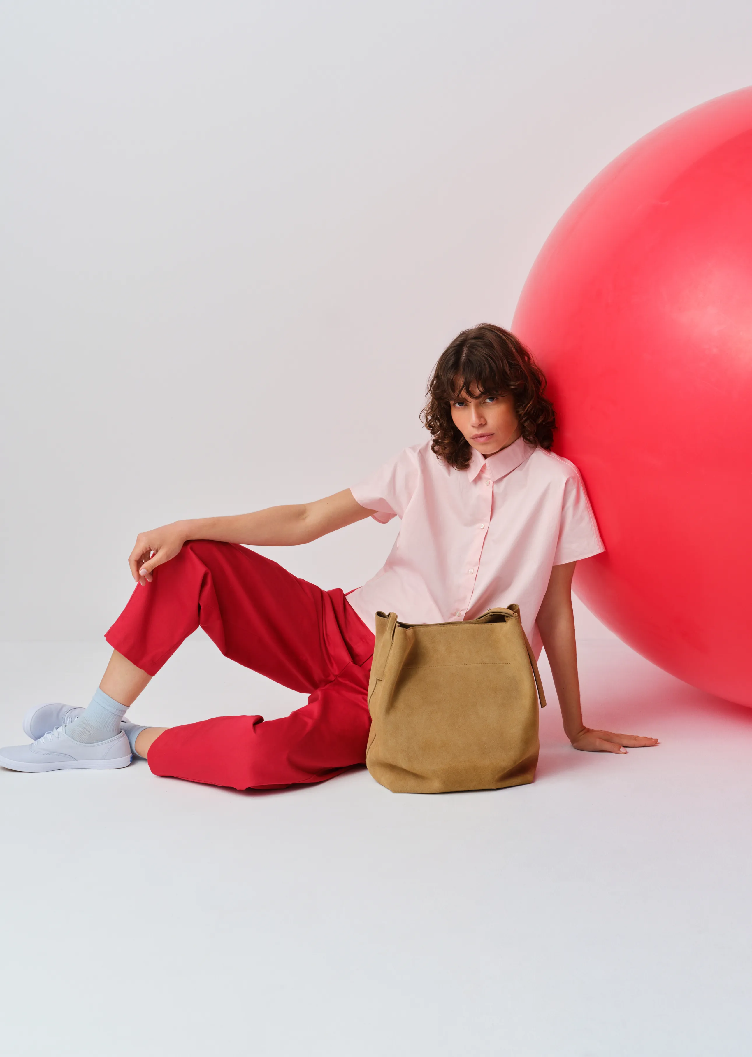 Model in pink shirt and red pants, sitting beside a large red ball.
