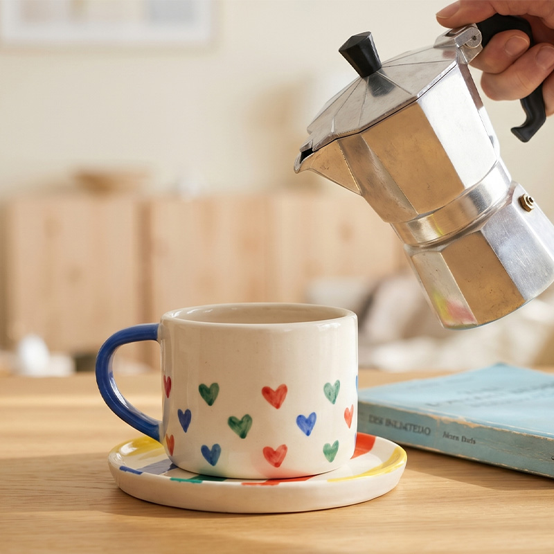A hand pouring coffee from a moka pot into a heart-patterned mug.
