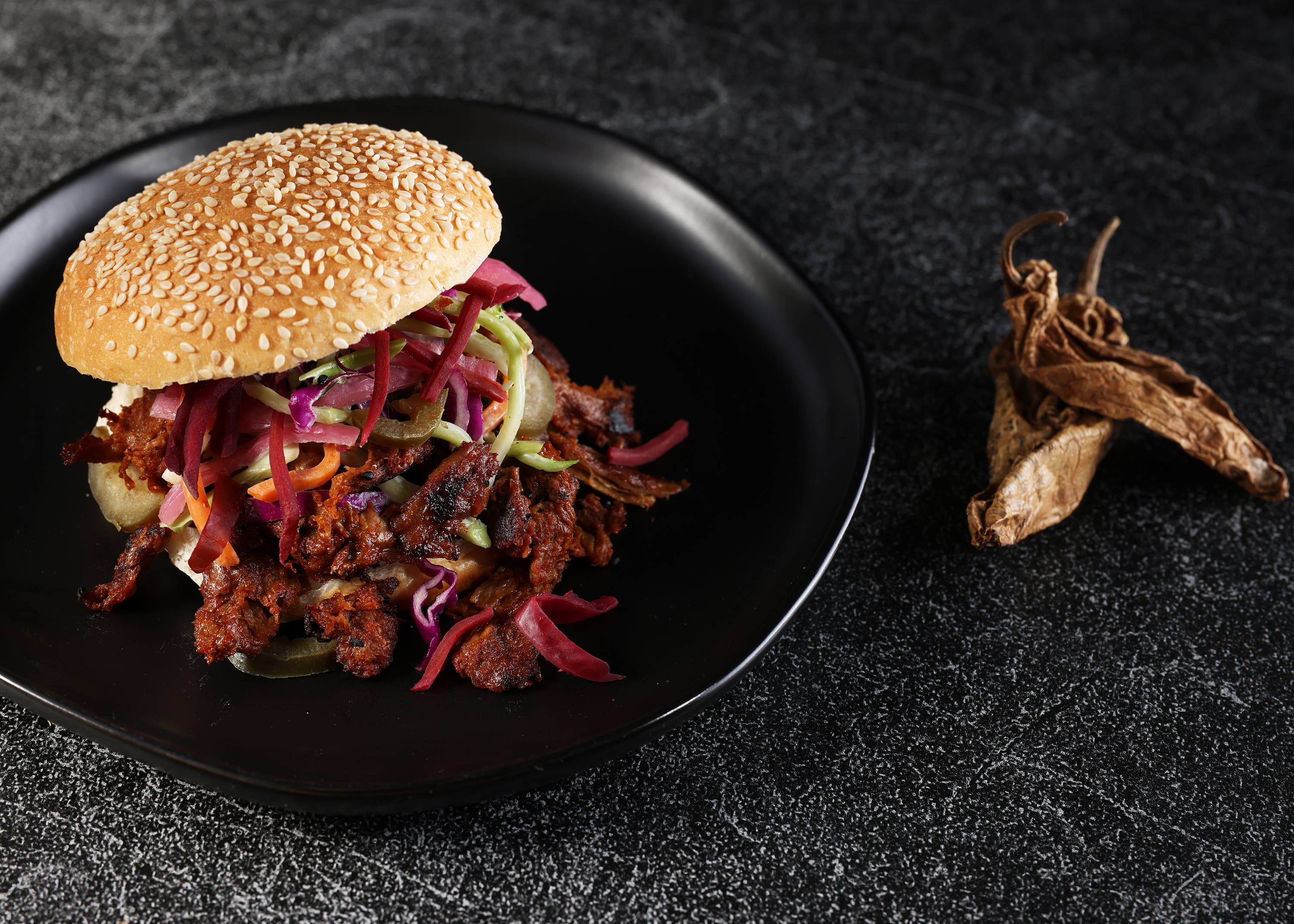 Texas BBQ Harvest Shreds on a bun with pickled red onions and slaw on a black plate next to dried chipotle peppers