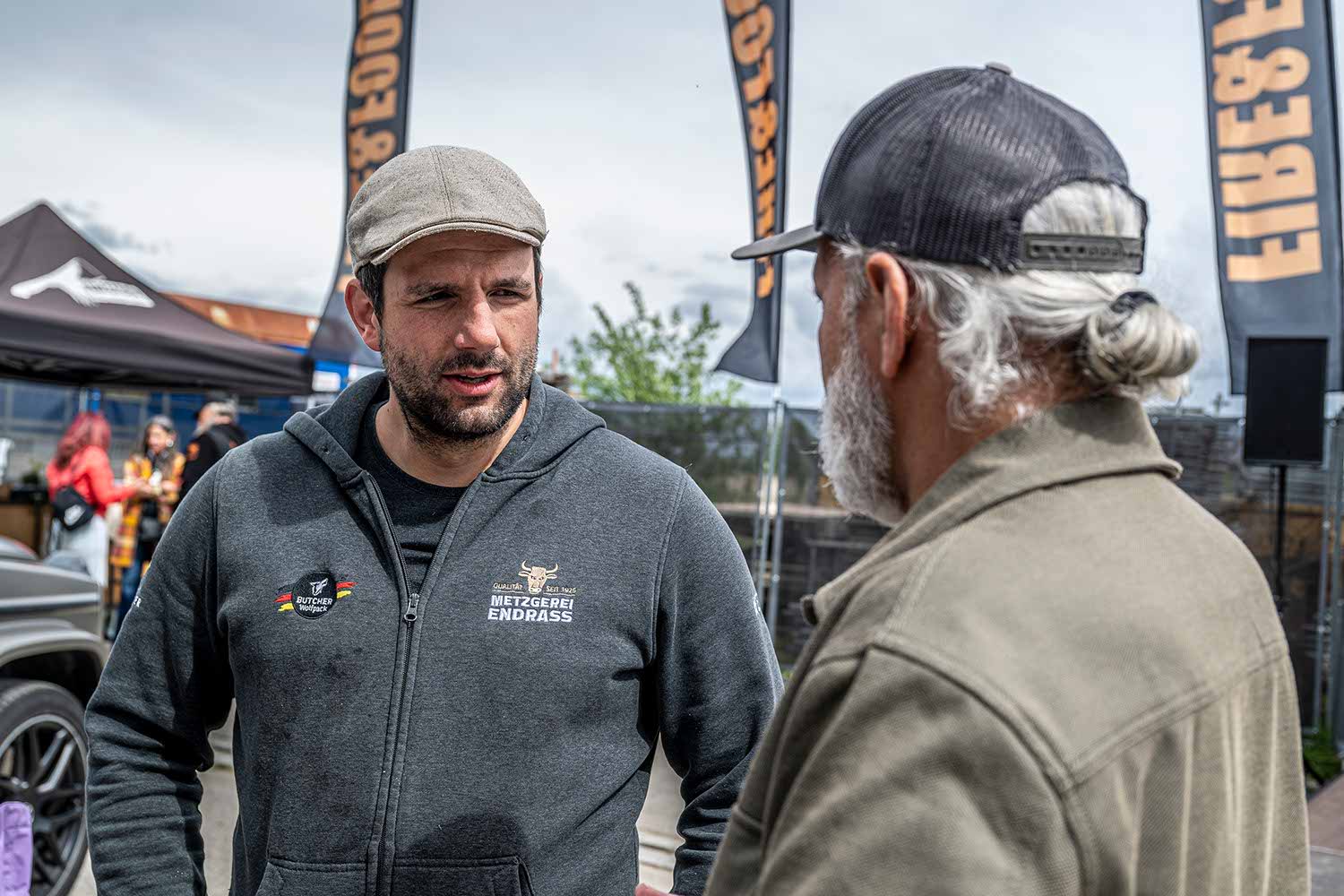Two men talking at an outdoor event with banners in the background.