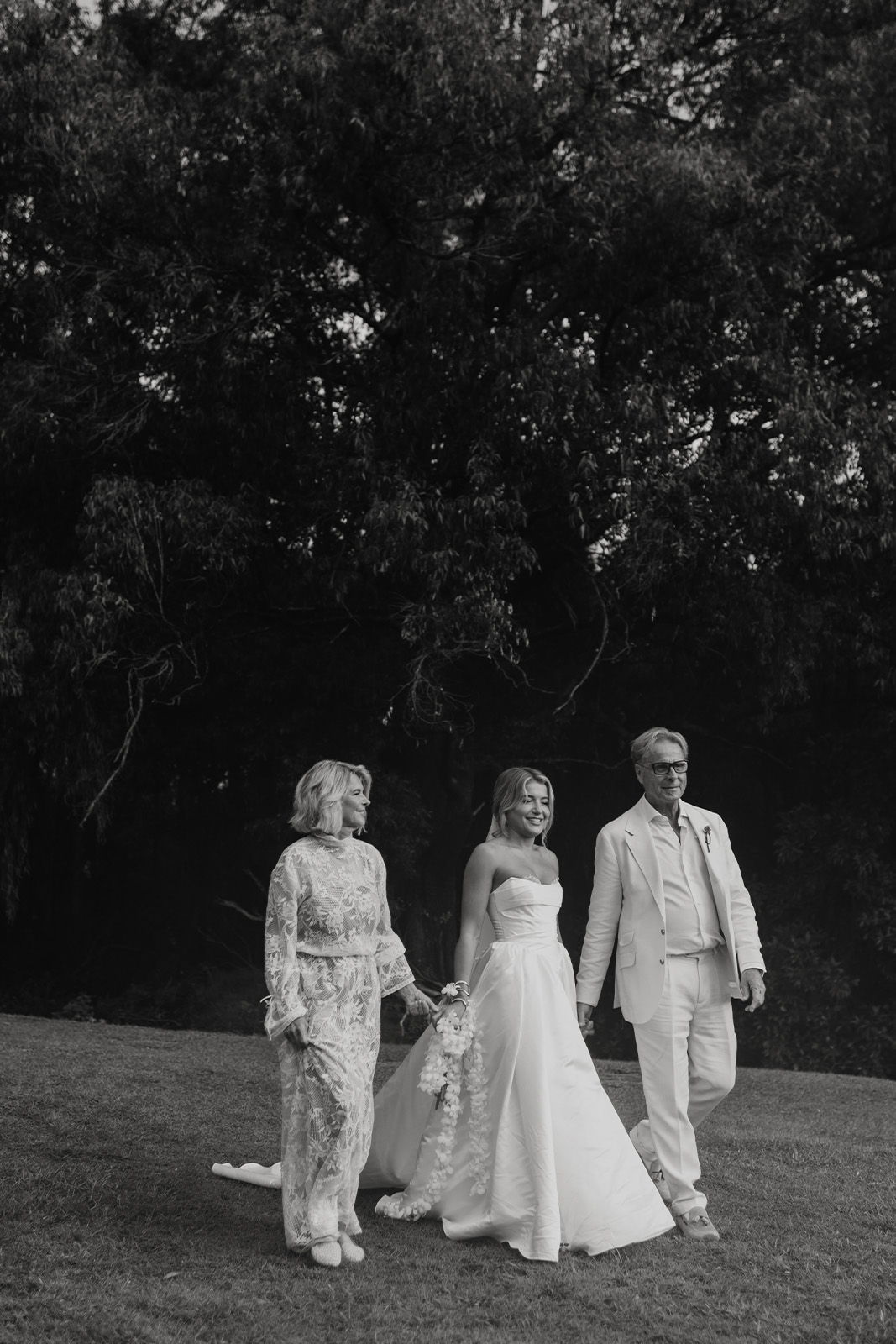 Bride walking with two people in formal attire, surrounded by greenery.