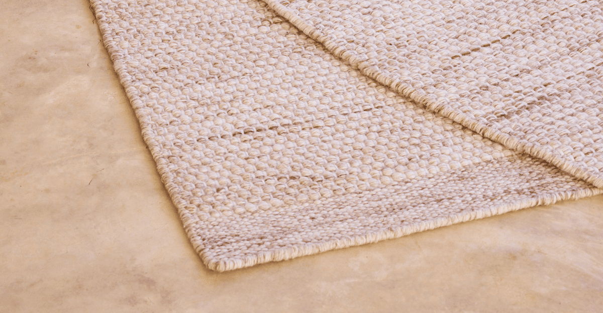 Close-up of a textured neutral outdoor rug corner on a stone patio surface, highlighting durable weave, low-profile construction, and material detail for patio use.