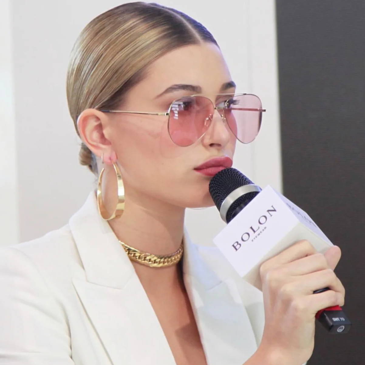 Close-up of a woman with slicked-back blonde hair wearing oversized pink-tinted aviator sunglasses, large gold hoop earrings, a chunky gold chain necklace, and a white blazer while speaking into a handheld microphone labeled 