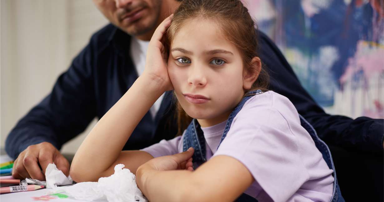 Girl leaning sadly on her elbow, doing homework with her dad – she has allergies and there are crumpled tissues on the table 