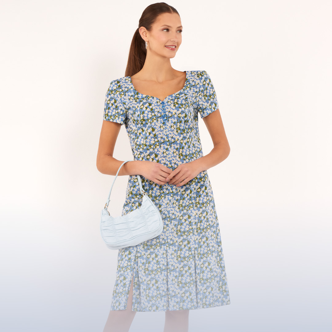 Woman in a floral dress holding a handbag, smiling at the camera.