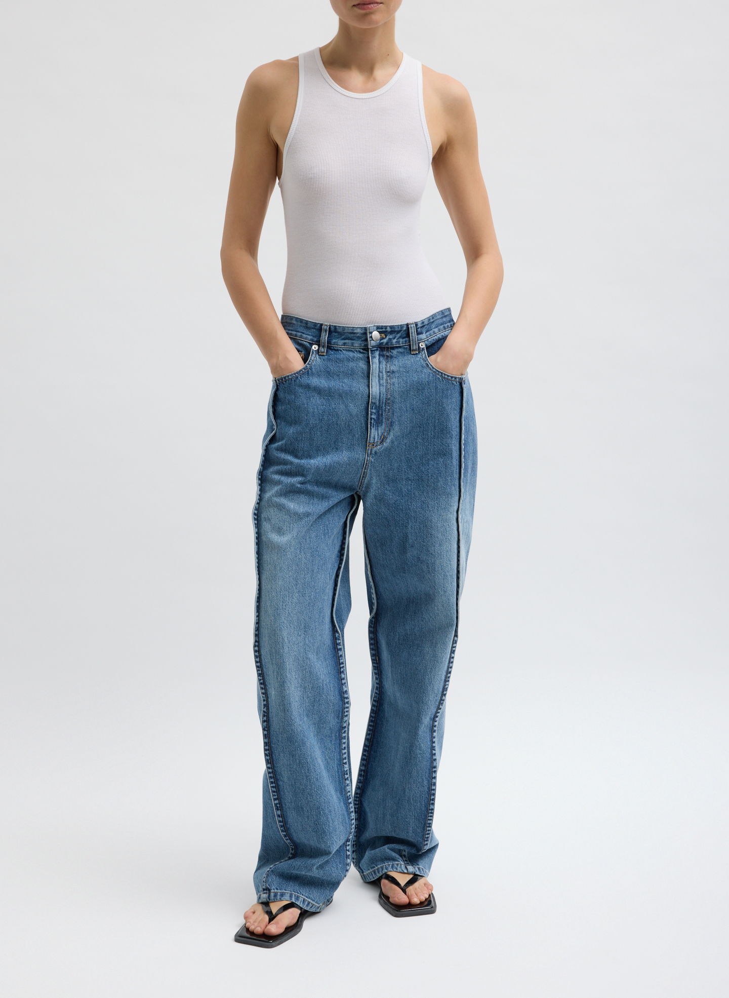 Model wearing a white tank top and blue jeans, hands in pockets.