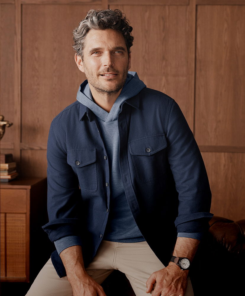 Man sitting against a wooden backdrop, wearing a navy jacket and hoodie.