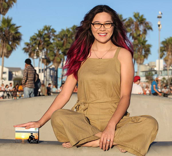 Asian woman with long red hair in tan jumpsuit sitting outside cross legged with right hand on box of Femmenessence MacaHarmony