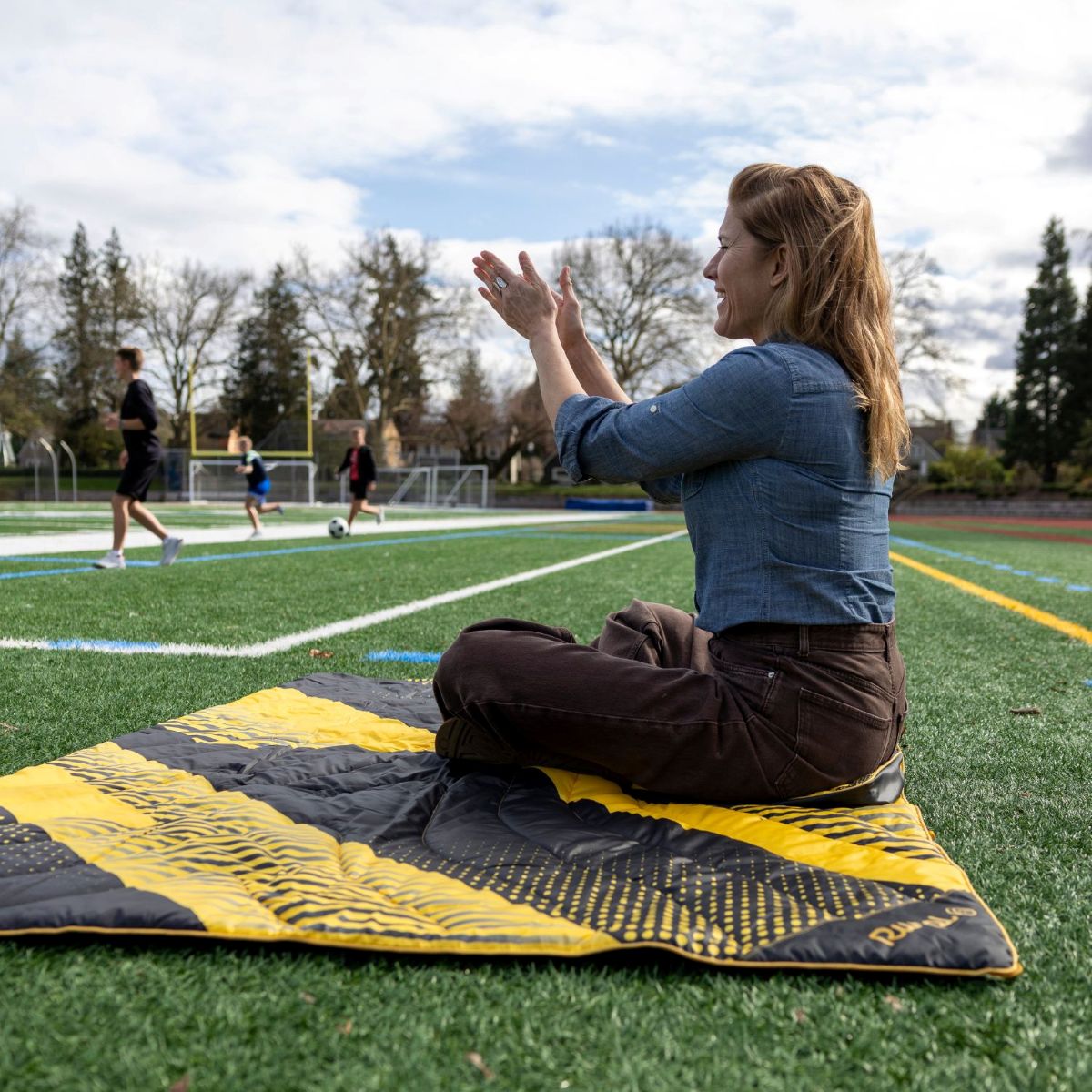 Woman sitting on a mat, clapping hands, with people playing in the background.