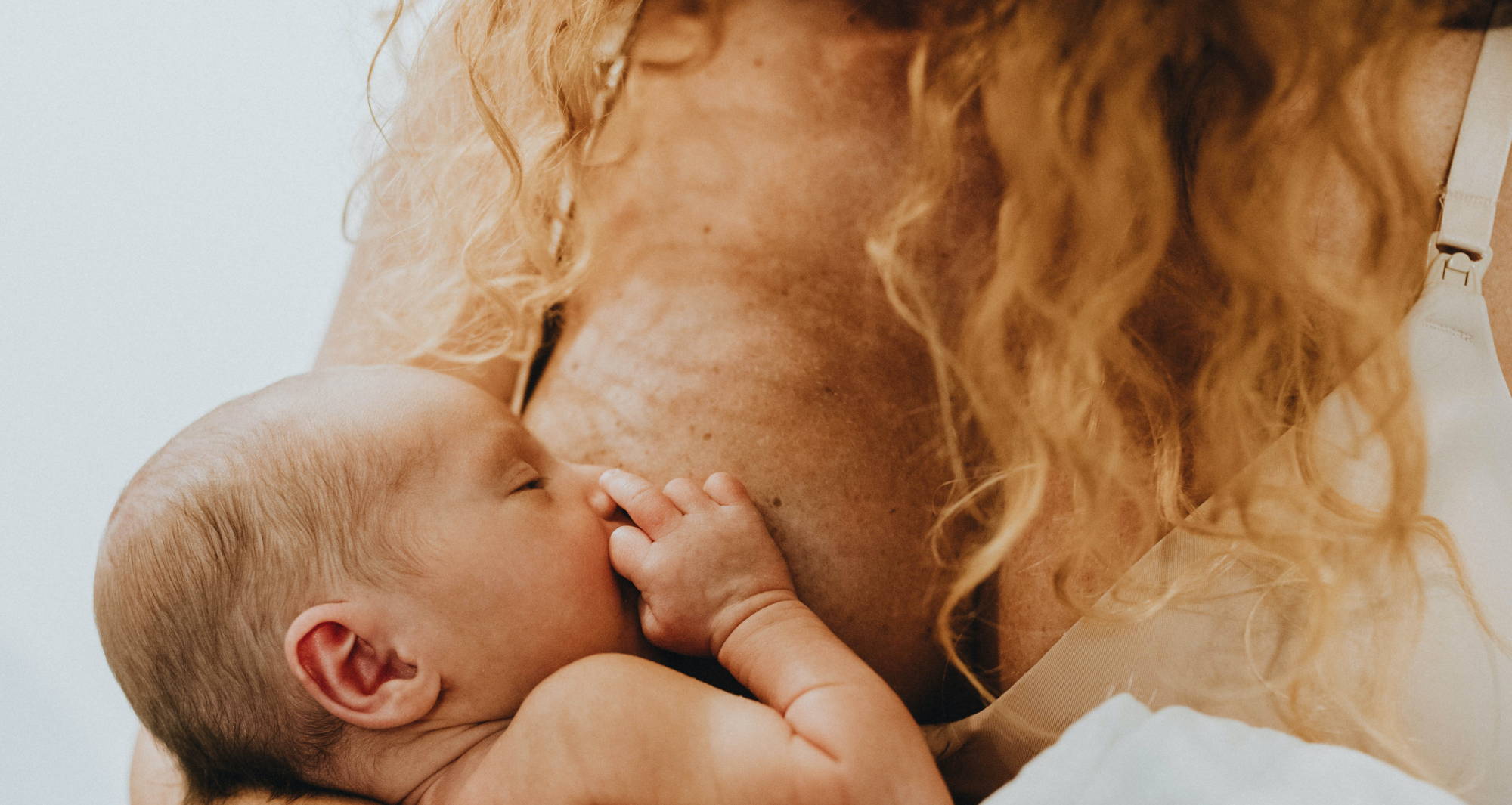  A woman with curly blond hair wearing a beige bra breastfeeds an infant.