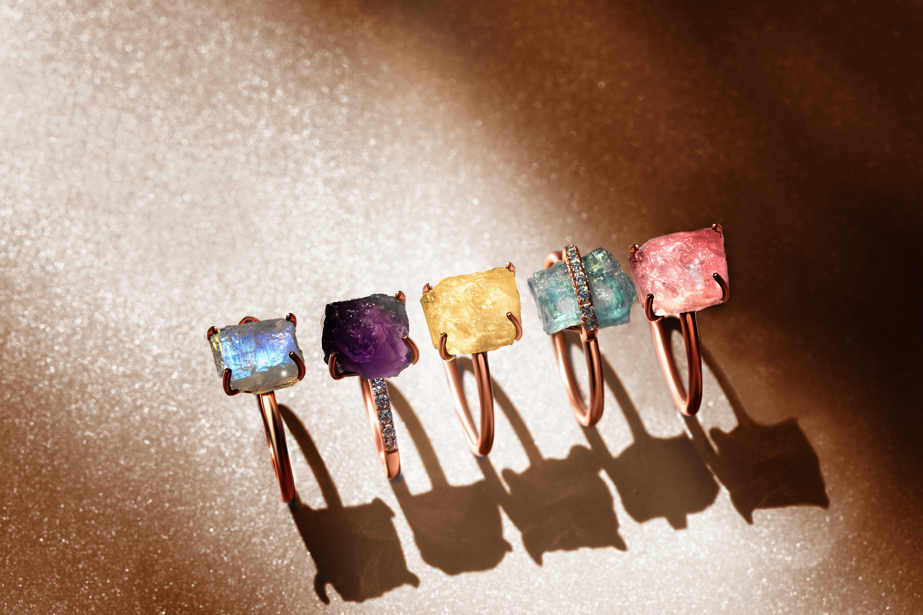 Five different Raw Crystal gemstone rings are presented on a golden surface.