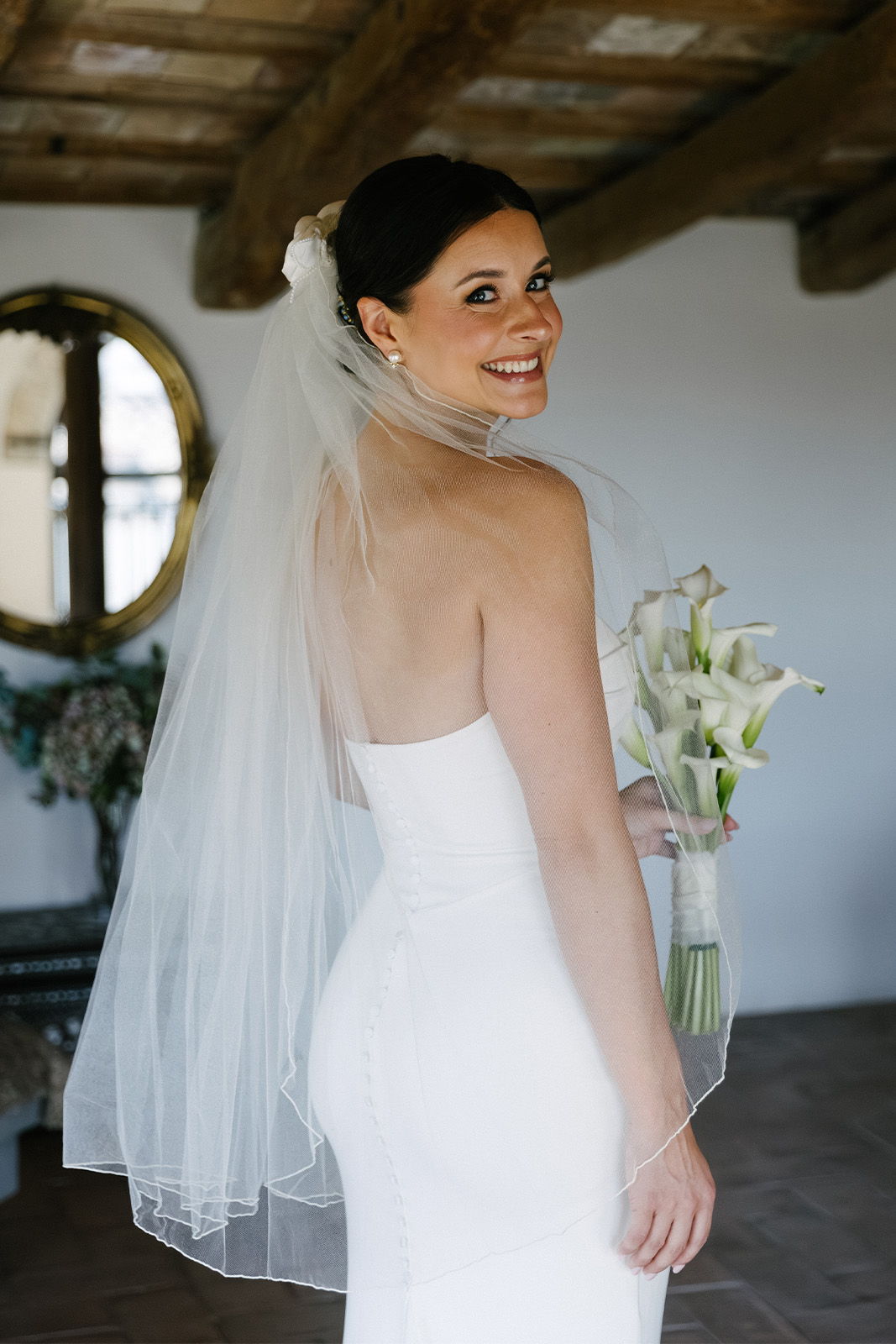 Bride in a white dress holding lilies, smiling over her shoulder.