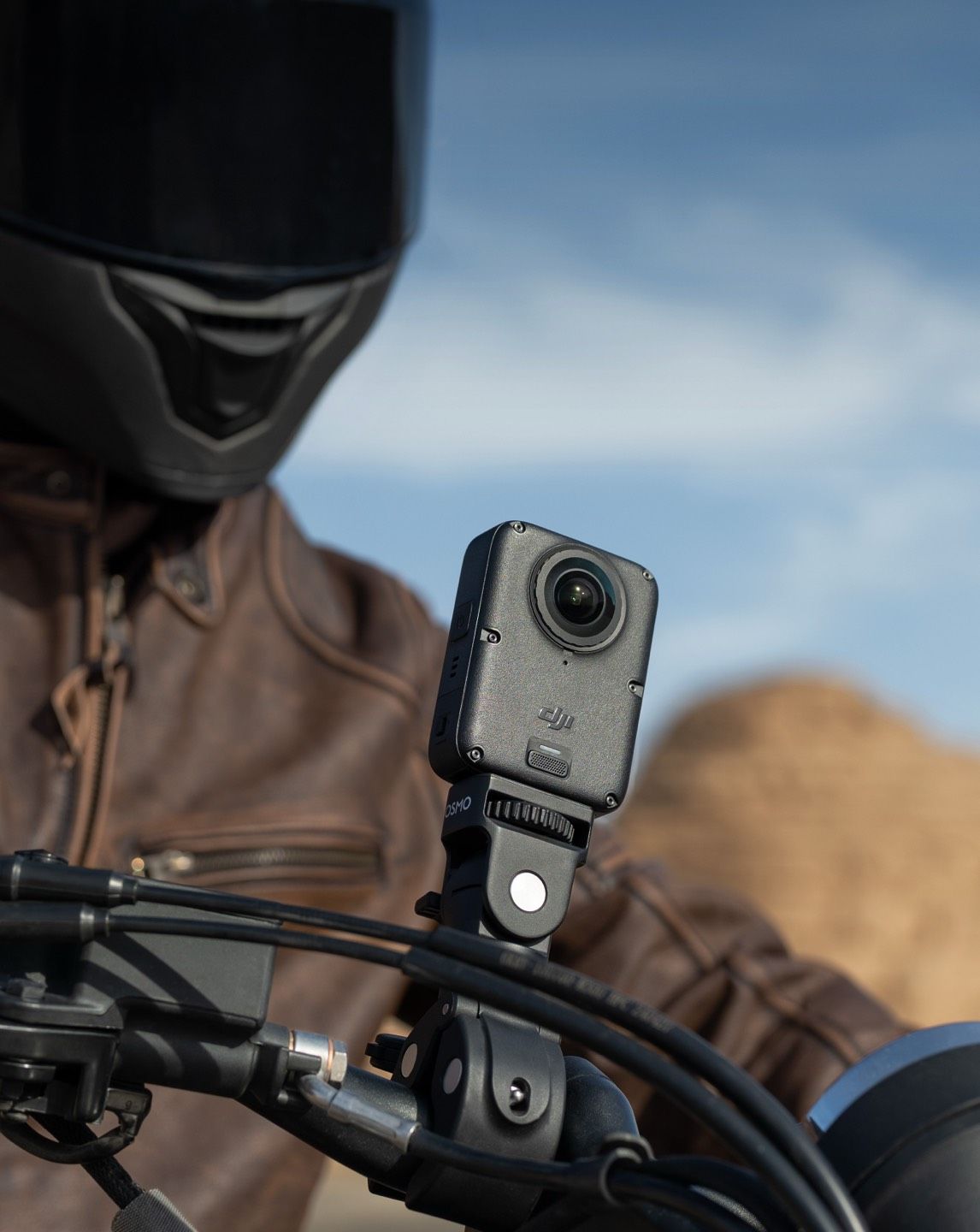A rider in a brown jacket with a camera mounted on a motorcycle handlebar.