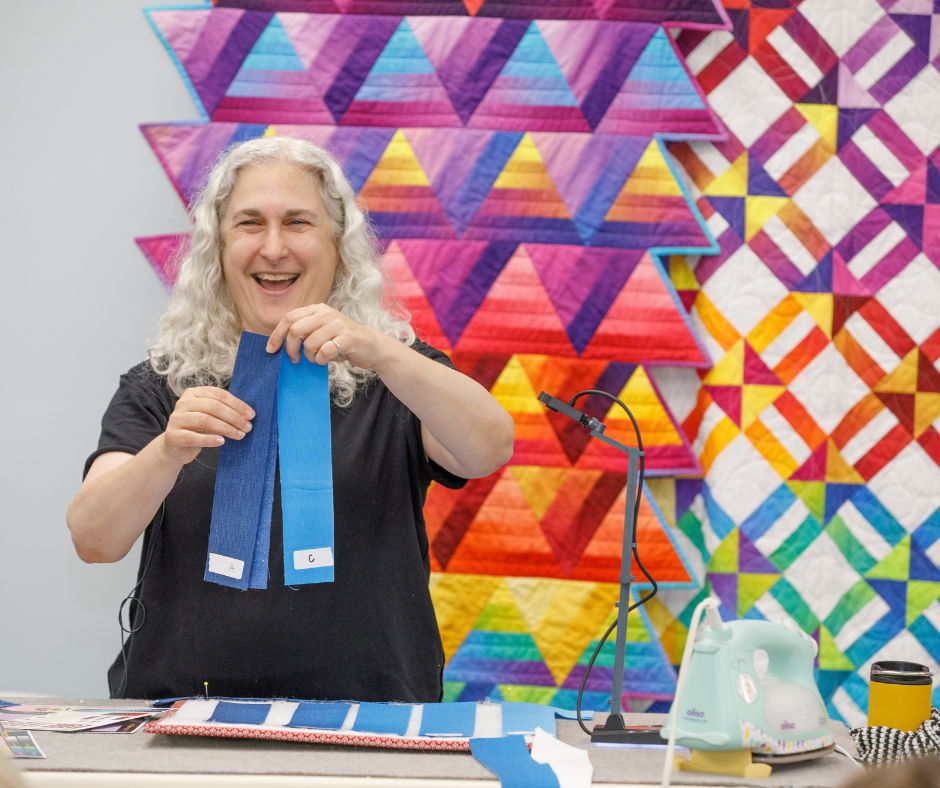 Person holding blue fabric strips in front of colorful quilt background.