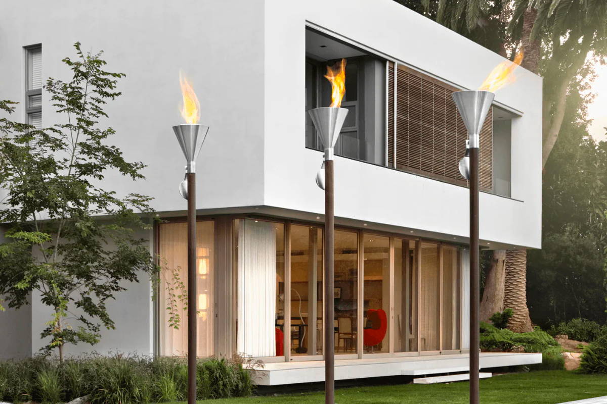 Flames dance out of the tops of three stainless steel garden torches in front of a modern white house. 