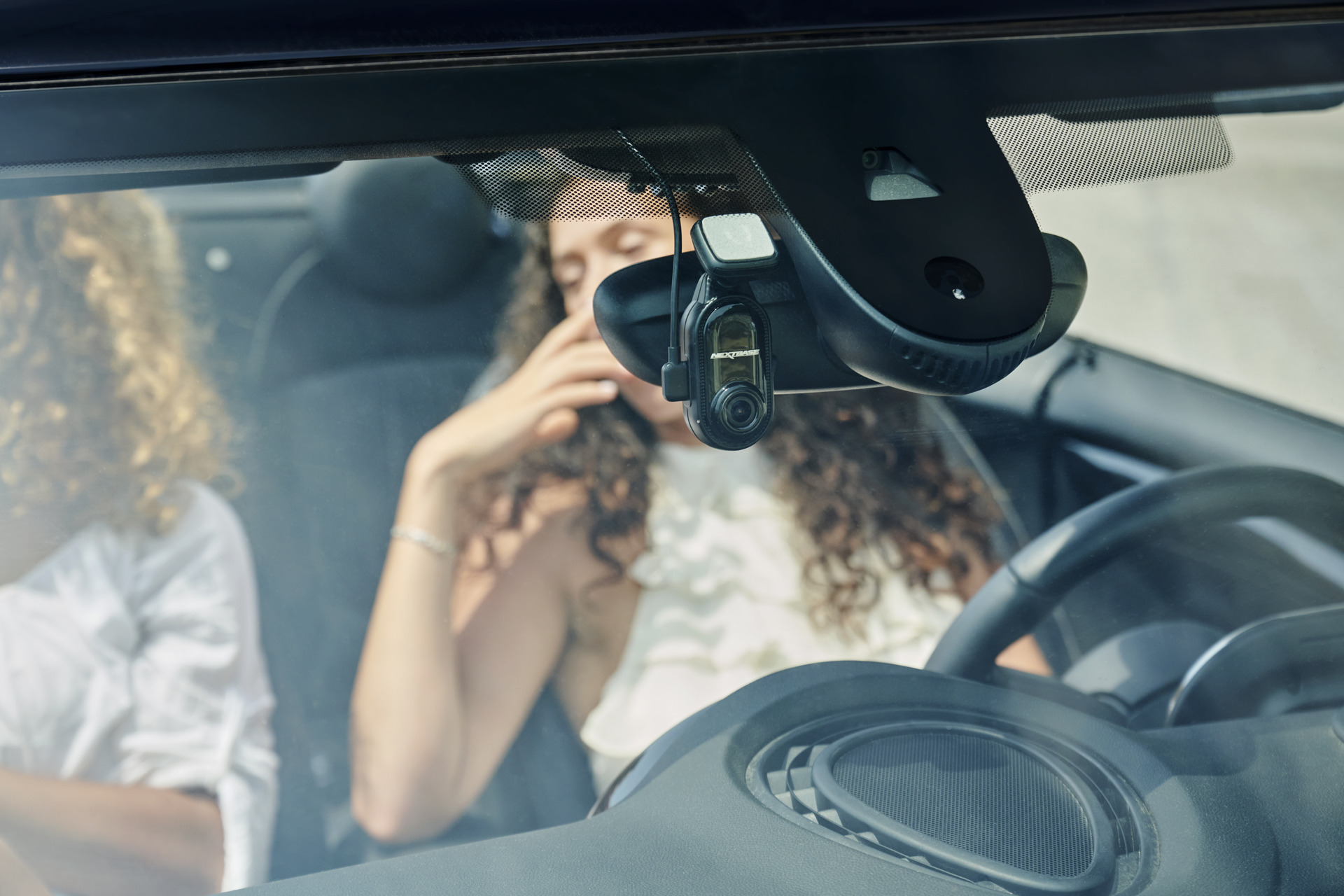 Nextbase Piqo dash cam mounted on a windshield with two women smiling in the background.