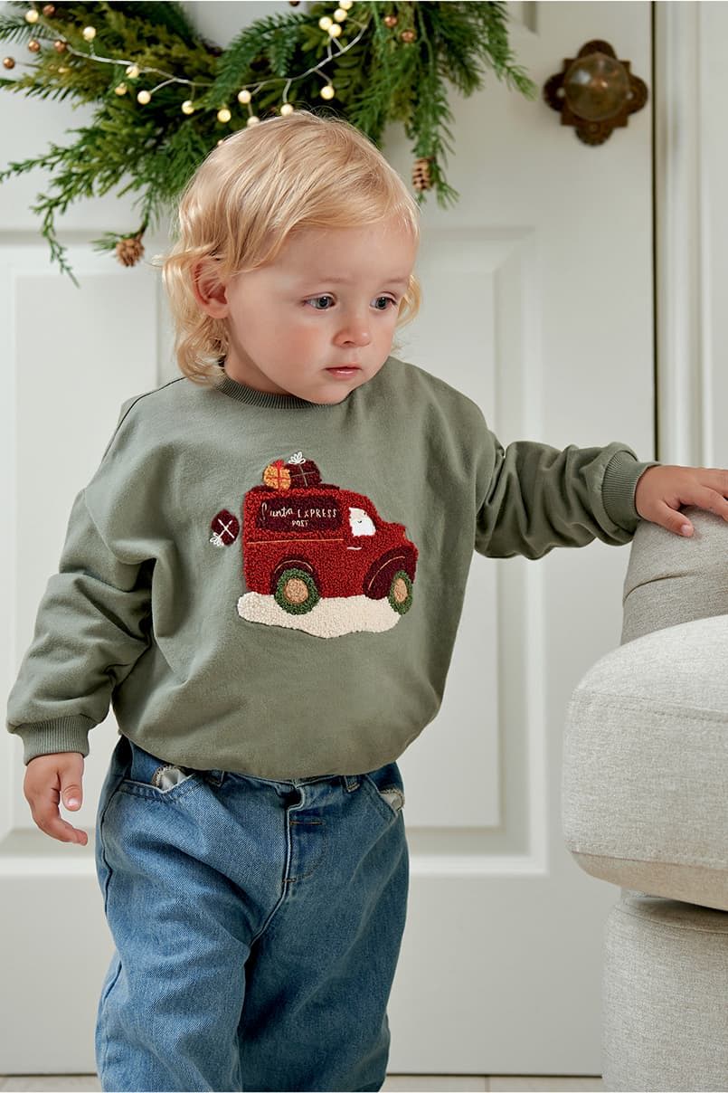 Toddler in green sweatshirt with truck design, walking near a couch.