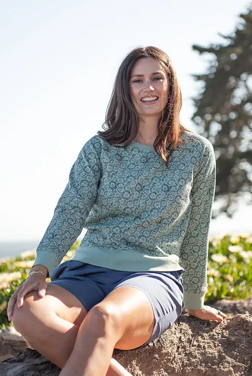 Smiling woman in patterned sweater sitting outdoors by the ocean.