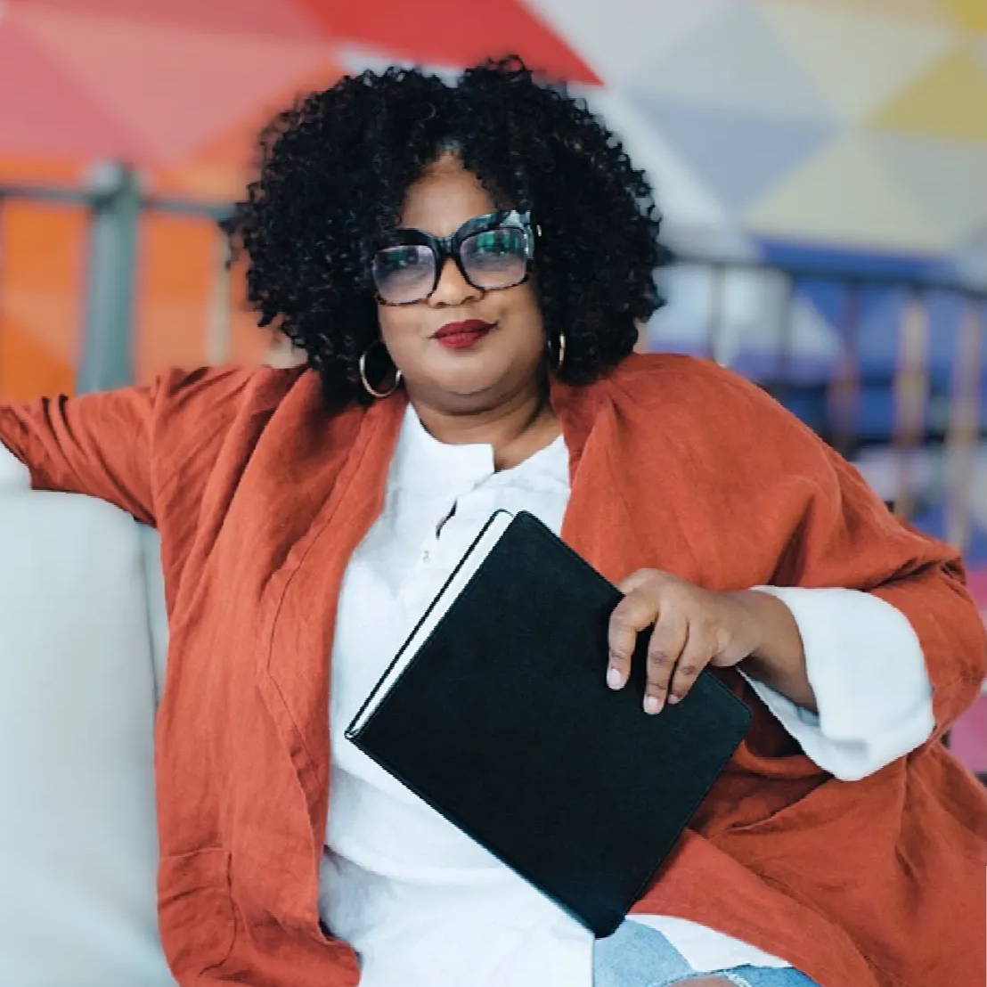 A woman relaxes on a couch hlding a book and looking straight into the camera