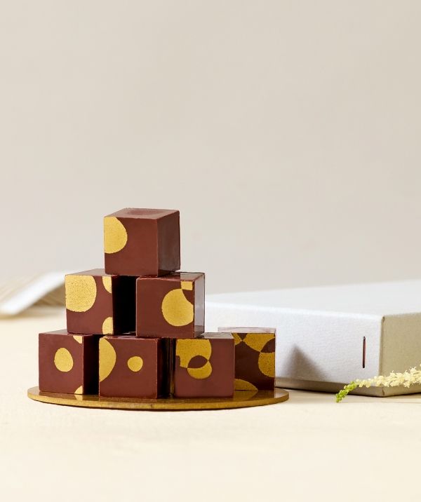 Chocolate cubes with golden dots, arranged on a plate beside a white box.
