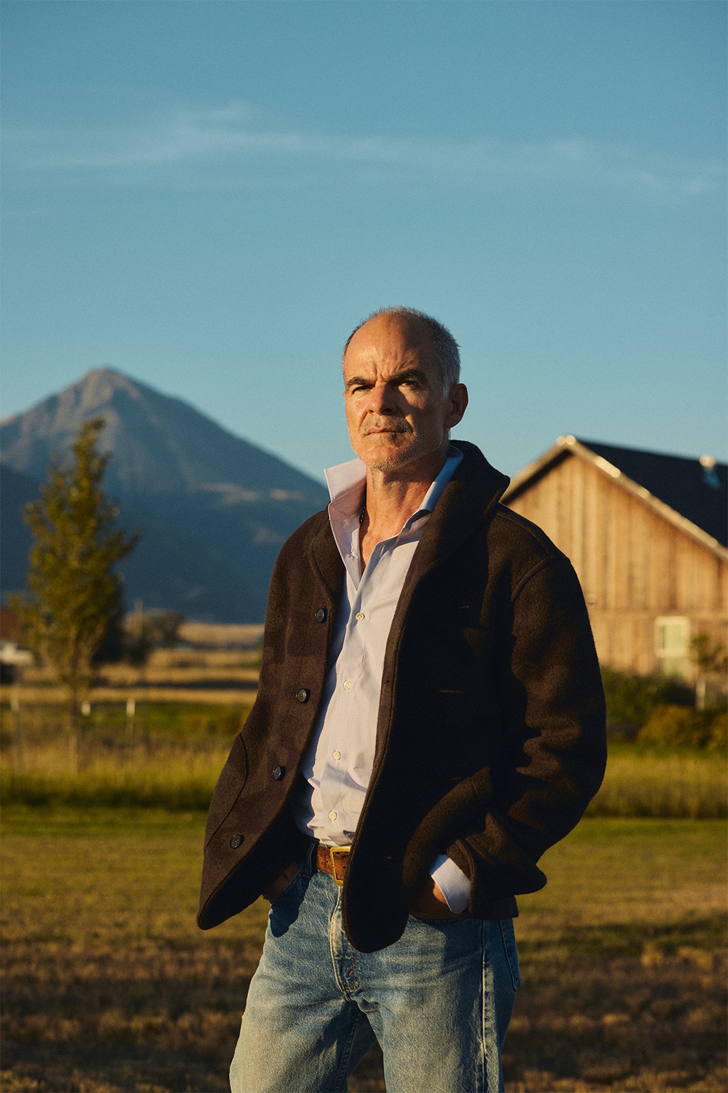 Micahel Kelly outside a barn in Pray Montana at sunrise wearing the Rivay Bridges Oxford Workshirt in Navy, the Hale Wool Cardigan in Olive Camouflage over the Corso Italian Dress Shirt in Classic Blue Micro Stripe and vintage pair of Levis with a mountain in the background