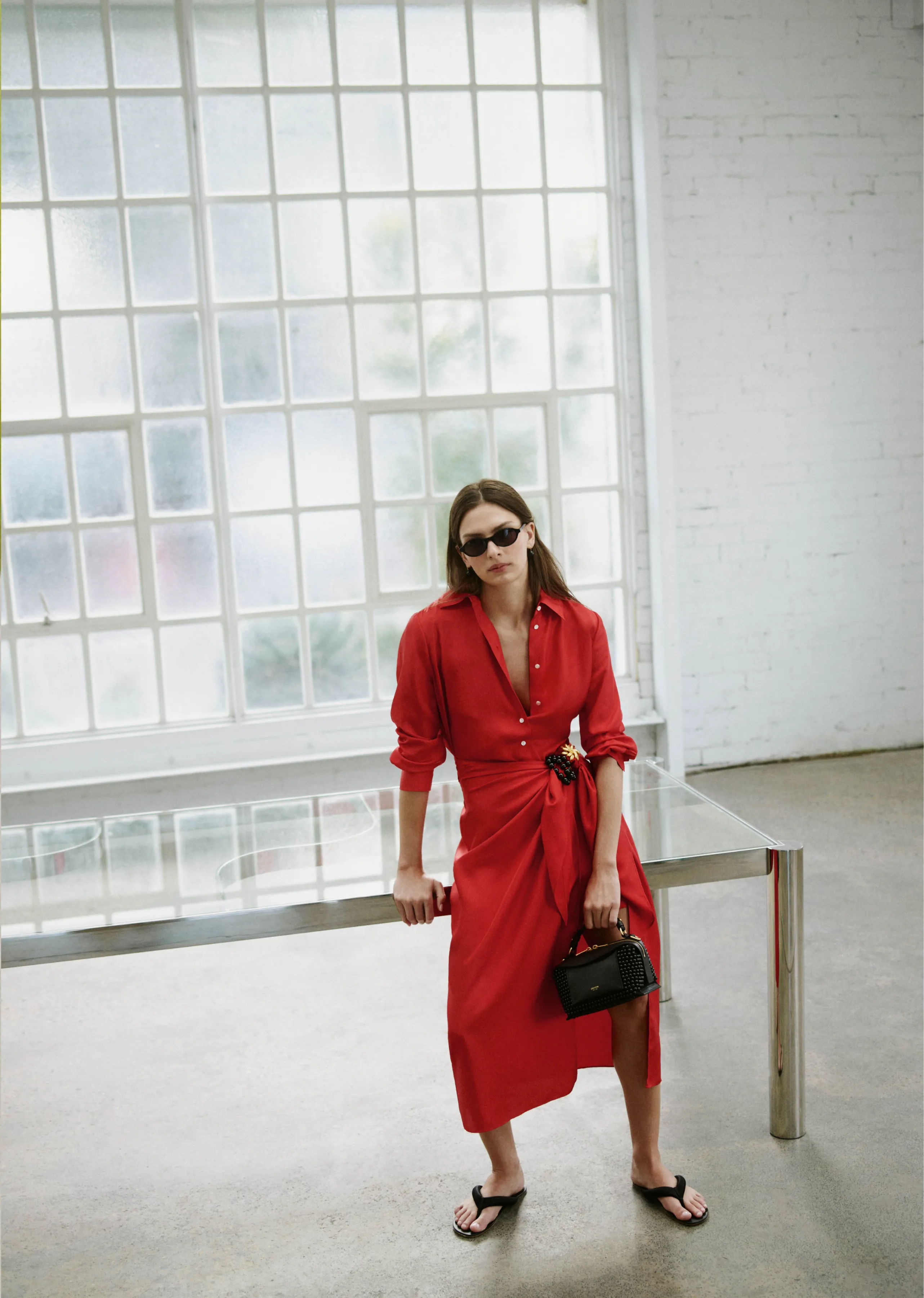 Woman in a red dress and sunglasses, standing in a bright, minimalistic space.