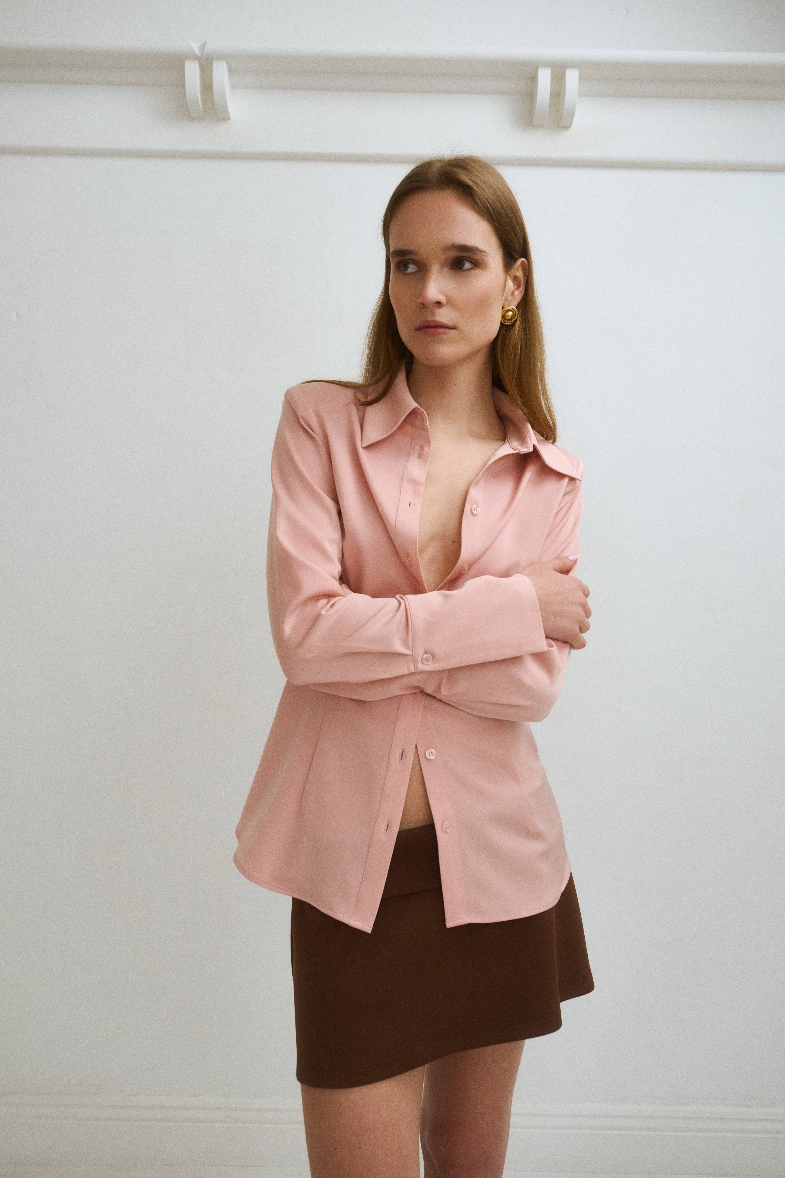 A woman in a pink shirt and brown skirt poses against a white wall.