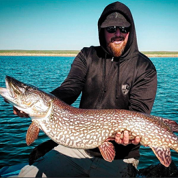 Mitch Dupuis standing by water holding up a large fish while wearing a black SA Company hooded performance shirt, sunglasses and a hat.