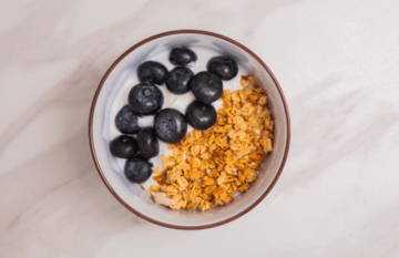Bowl of yogurt topped with blueberries and granola on a marble surface.
