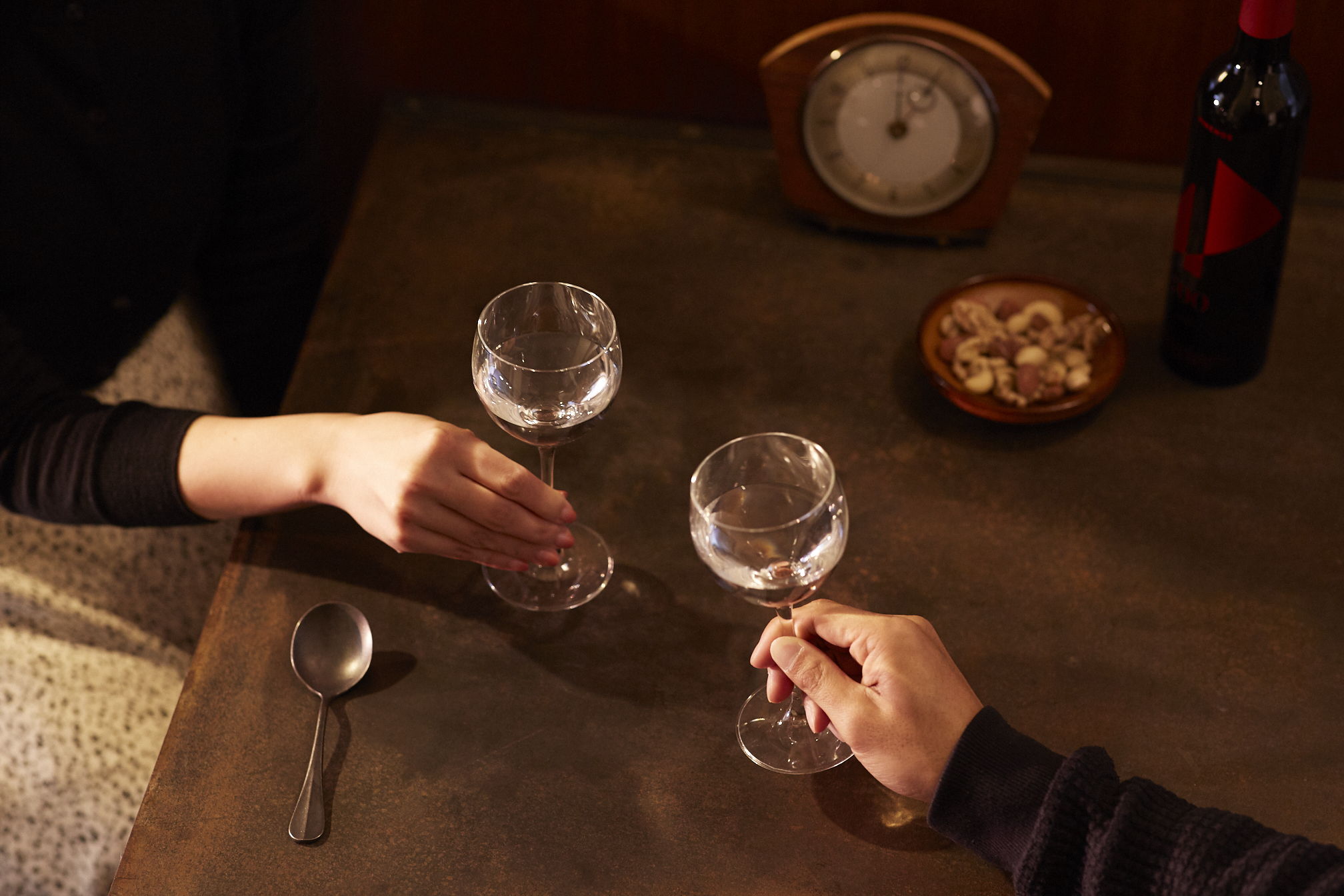 Hands toasting with glasses over a table with snacks and a clock.